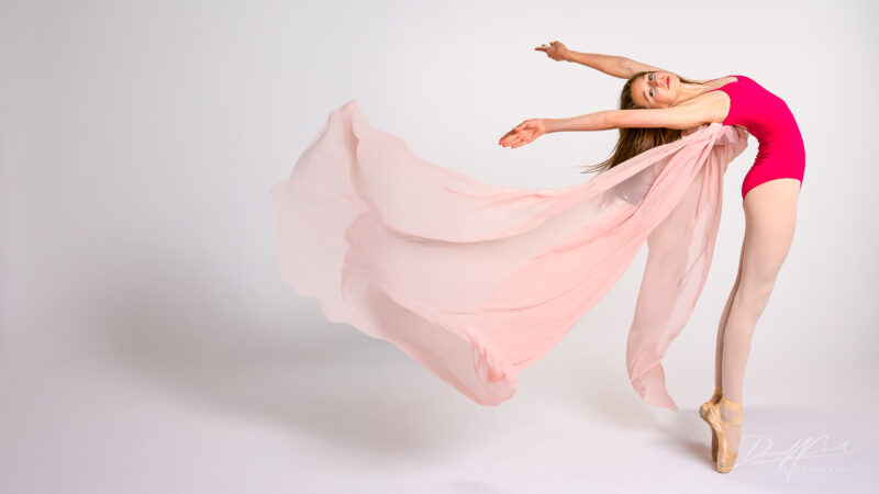 A graceful ballerina captured mid-leap during a studio photoshoot, wearing a vibrant pink leotard and pointe shoes. Her sheer, flowing fabric trails behind her, creating an ethereal sense of motion and elegance.