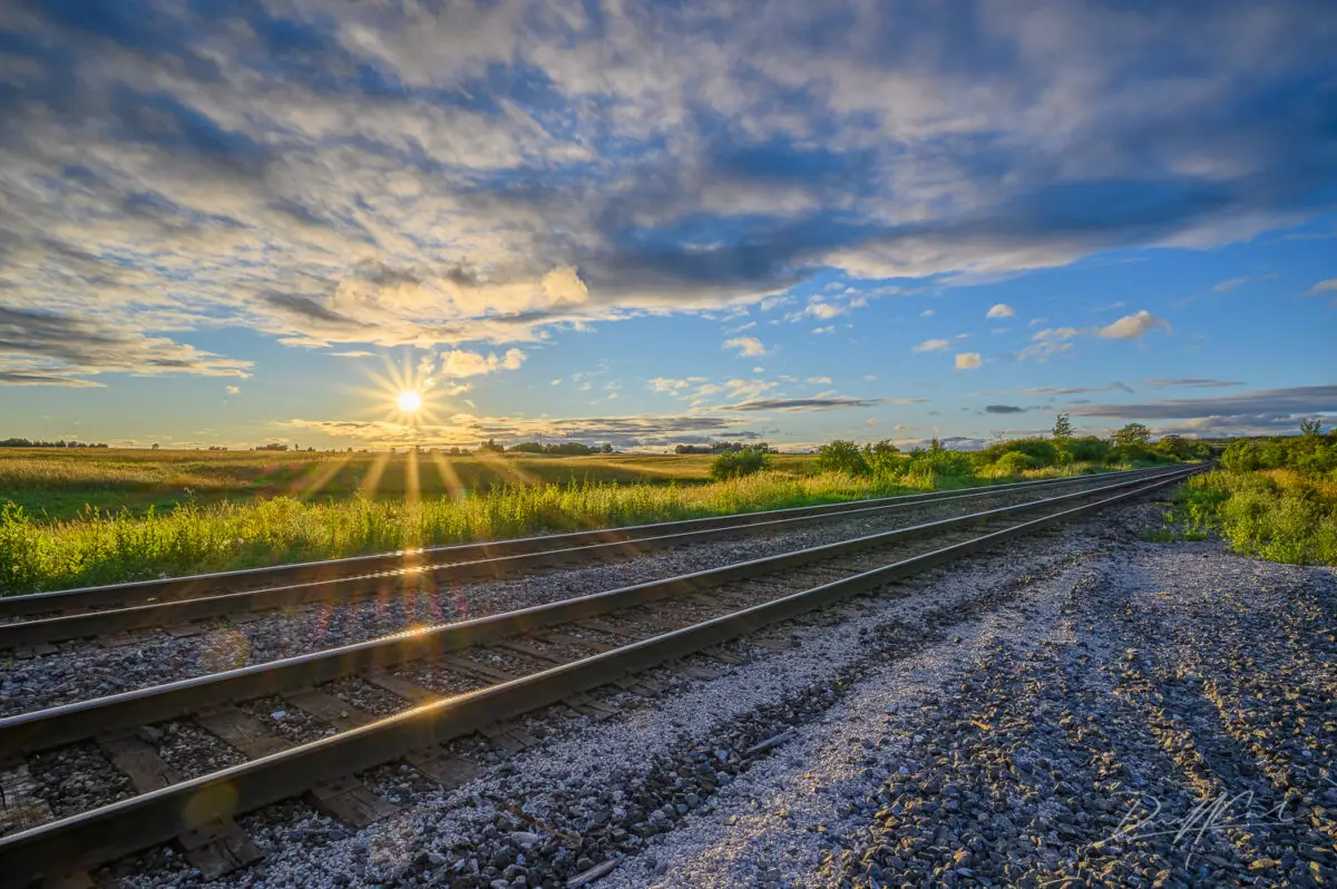 A warm July evening in Burlington, Ontario, with the sun setting low over quiet train tracks and open farmland. The golden light, scattered clouds, and long shadows create a peaceful scene with a stillness that invites you to pause and take it in. Everything lines up just right: the light, the land, and the sky.