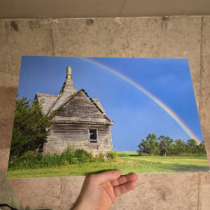 A vivid rainbow arching over a weathered abandoned wooden building surrounded by prairie grass and trees in Alberta.