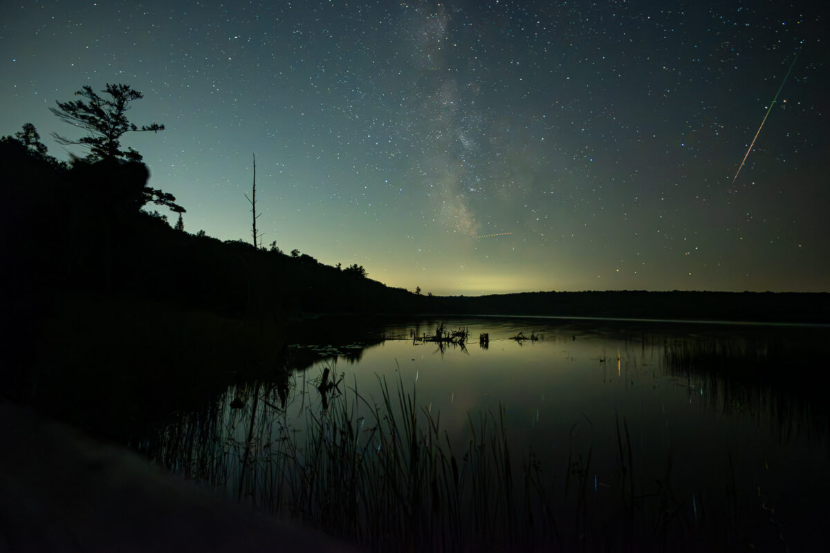 A Perseid meteor and the Milky Way above a calm lake at Awenda Provincial Park, reflected in the water during the 2026 meteor shower.